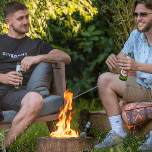 People drinking outside in the garden around El Fuego Fire Pit, the UK's most eco-friendly fire pit. A large flame comes out of the fire pit from the eco-friendly candle, giving a warm glow to the people.