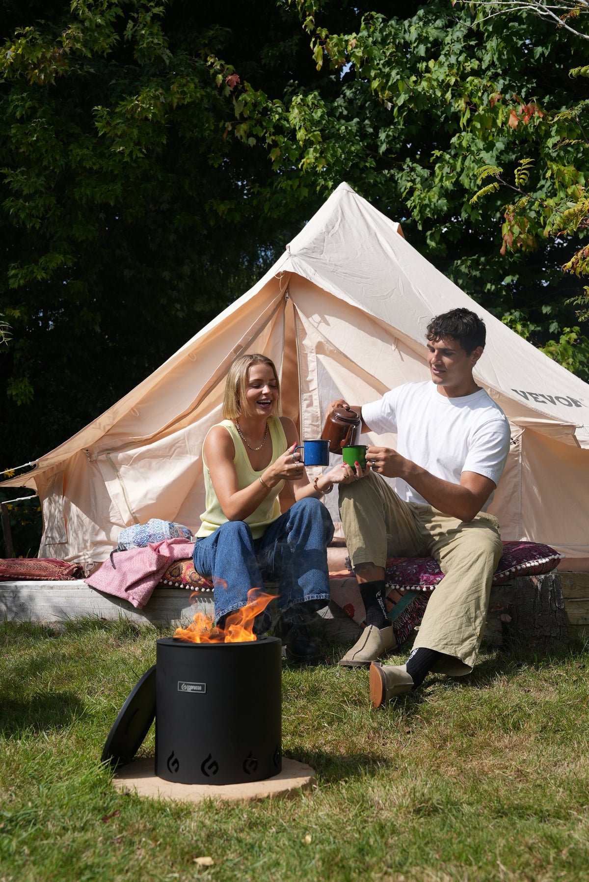 Two people sitting outside a canvas tent with mugs, an ECOFUEGO El Campo burning on the grass in front of them