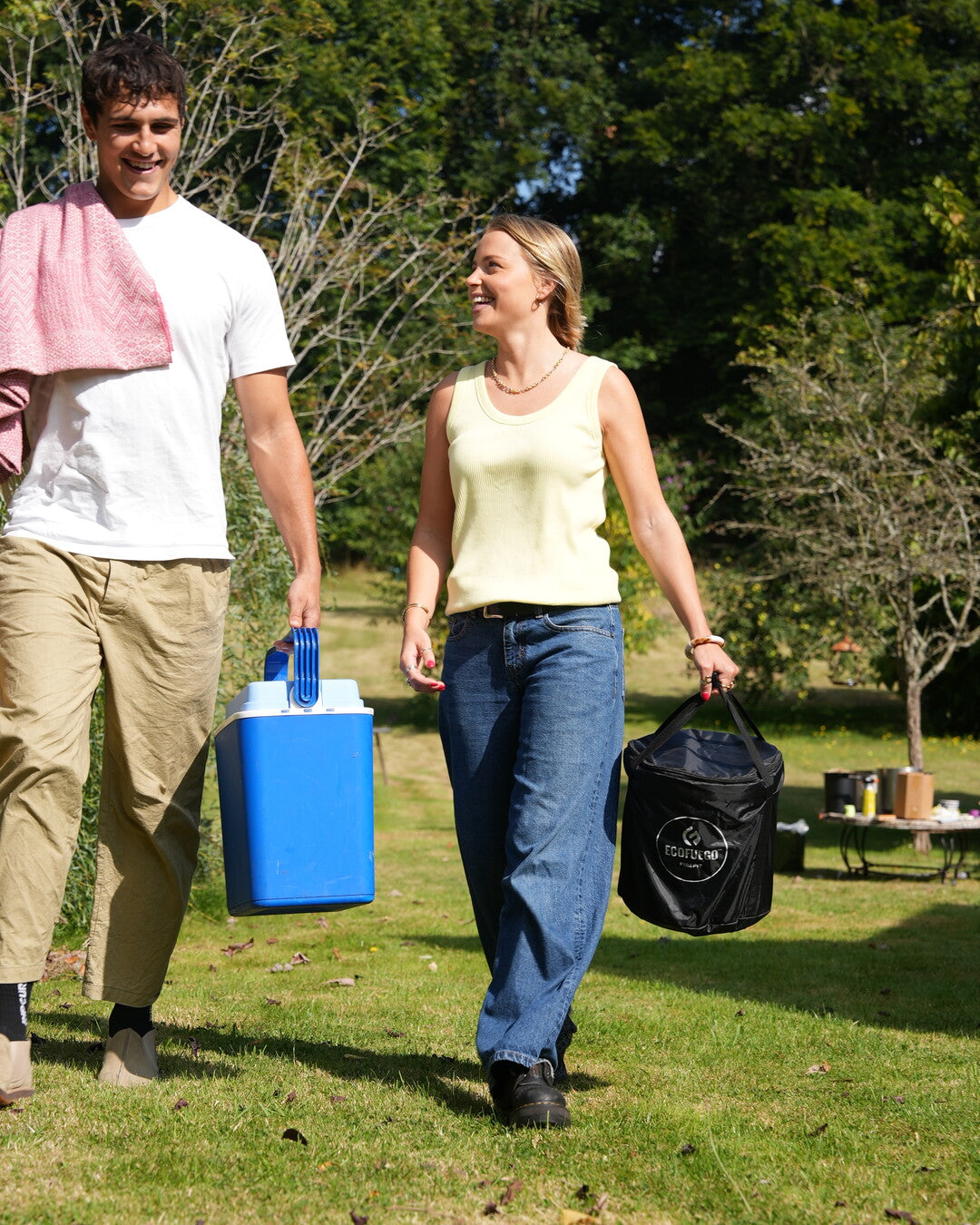 Two people walking across grass to a campground, one carrying an ECOFUEGO El Campo in its black travel case and the other a cooler
