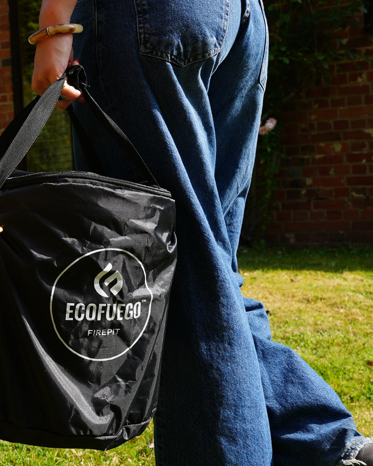 Close view of a person walking across grass with an ECOFUEGO El Campo in its black carry case hanging from one hand